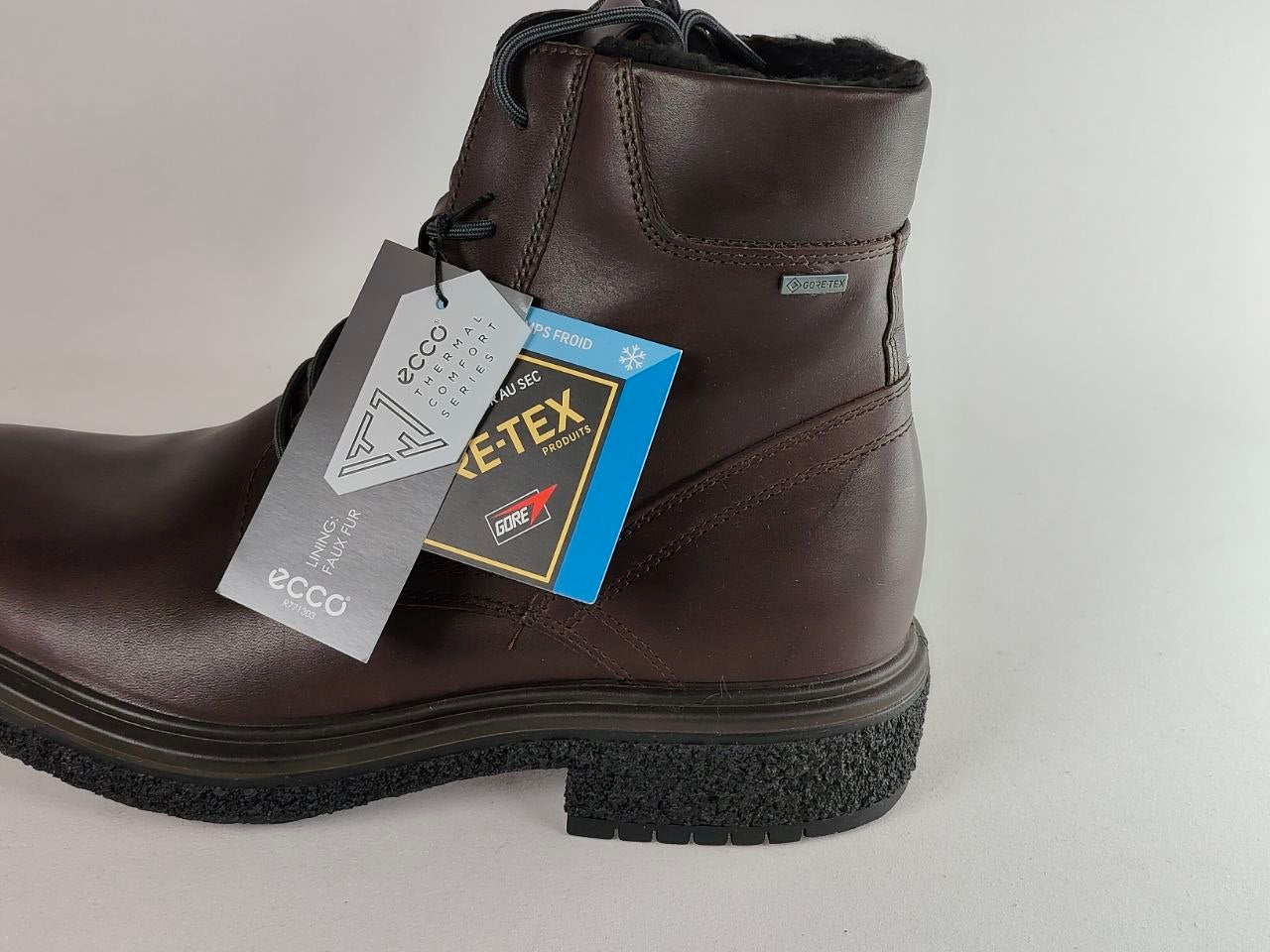 A dark brown leather ankle boot with black laces and a black textured sole, featuring a gray and blue Eccotox GORE-TEX waterproof tag attached, set against a plain light gray background.
