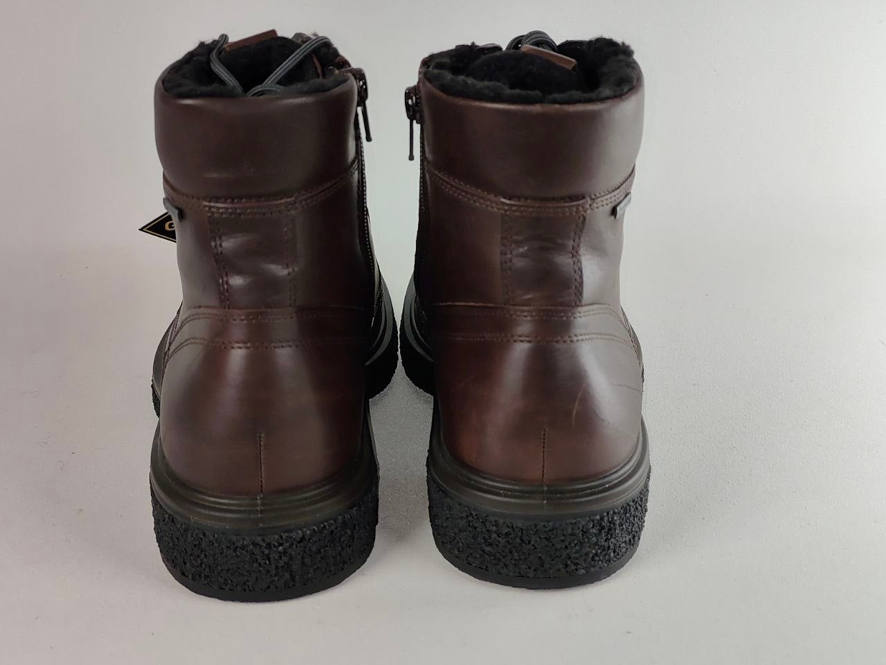 A pair of dark brown leather ankle boots with black textured soles and a black faux-fur lining, shown from the back against a light gray background.