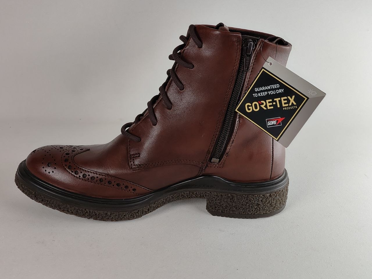 A brown leather lace-up ankle boot with a black side zipper and a hanging GORE-TEX tag, photographed on a light gray background.