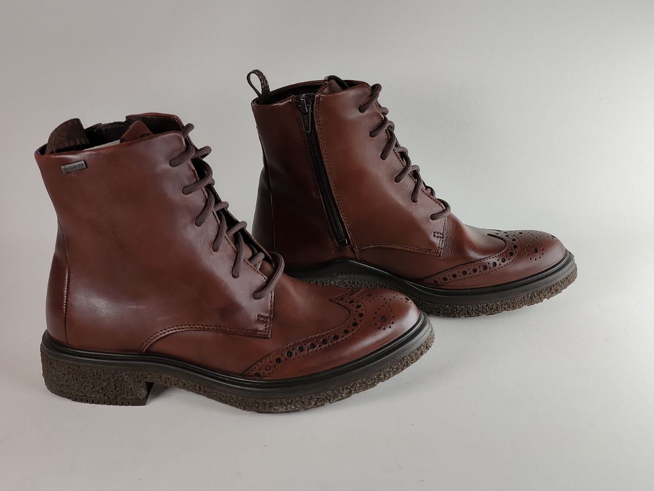 A pair of brown leather lace-up ankle boots with brogue detailing on the toe and a side zipper, photographed on a neutral light gray background.