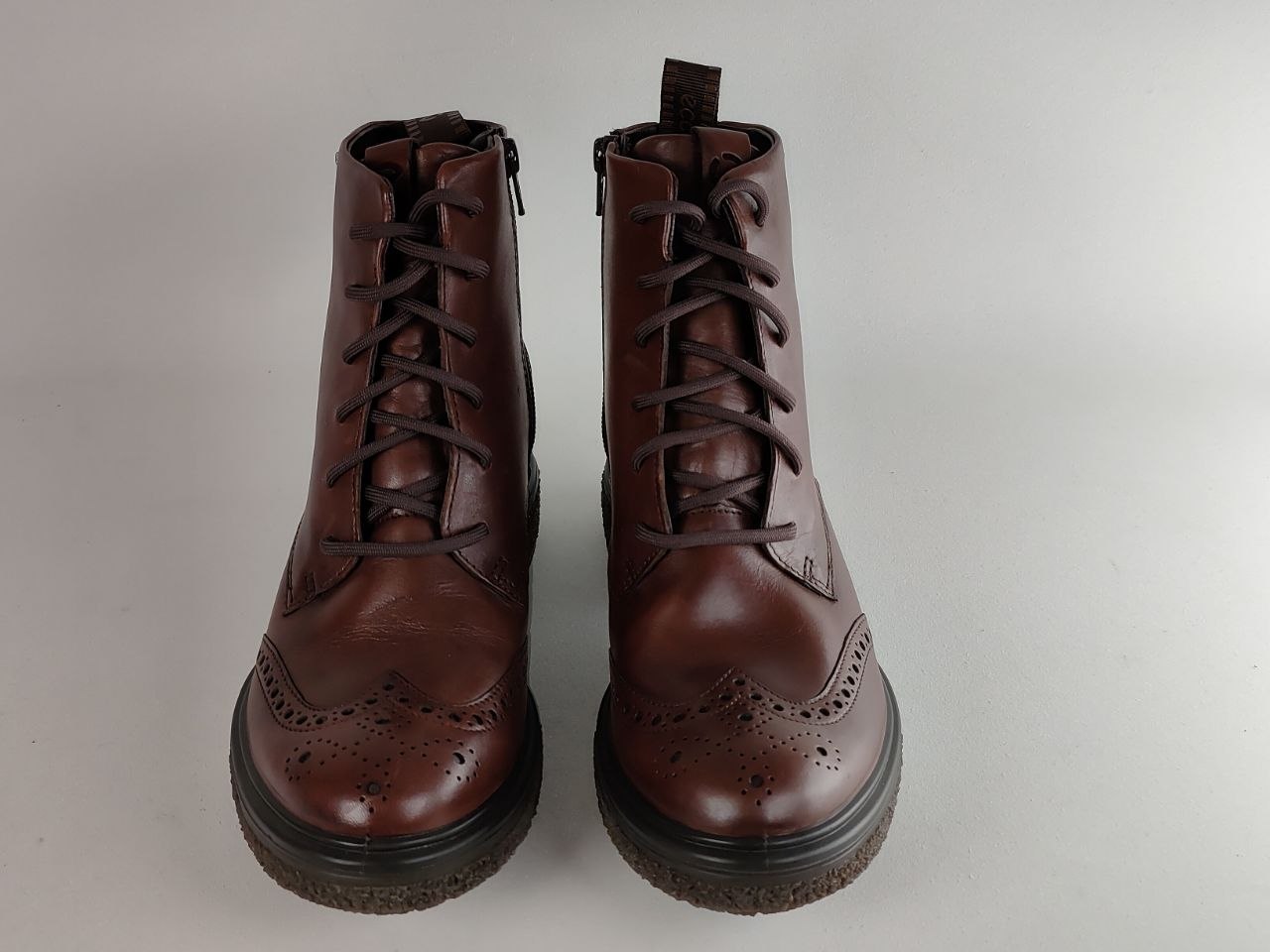 A pair of dark brown leather lace-up ankle boots with brogue detailing on the toes, photographed against a neutral light gray background.