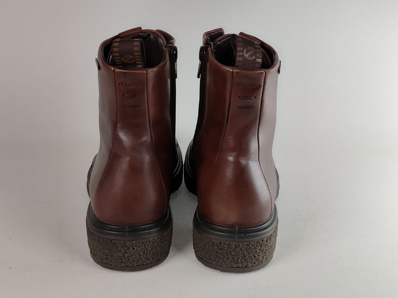A pair of brown leather ankle boots shown from the back, featuring dark textured soles, photographed against a neutral light gray background.