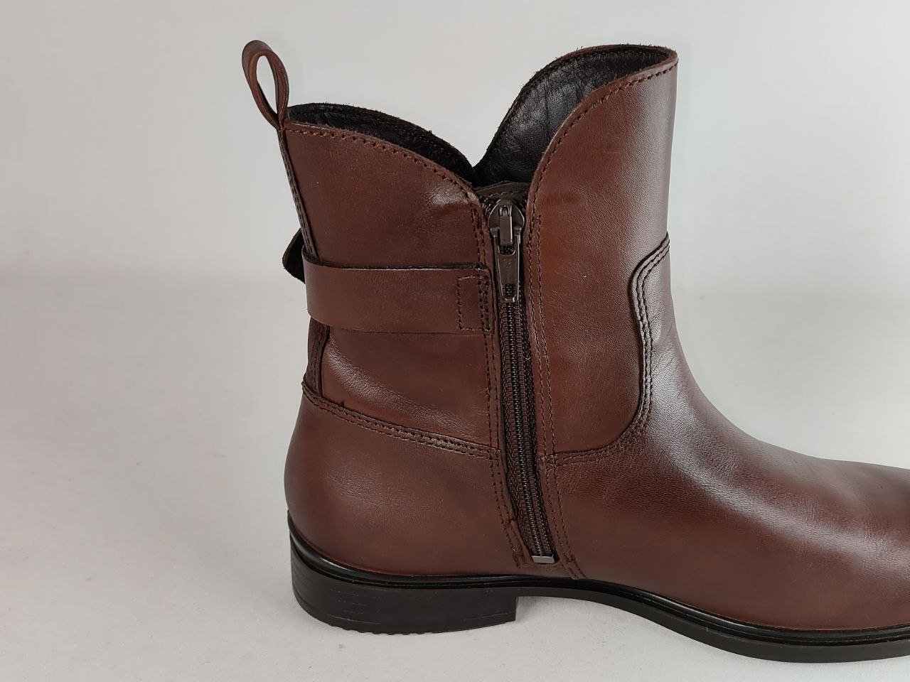 A brown leather ankle boot with a side zipper, elastic side strap, and pull tab, shown from a side profile view against a light gray background.