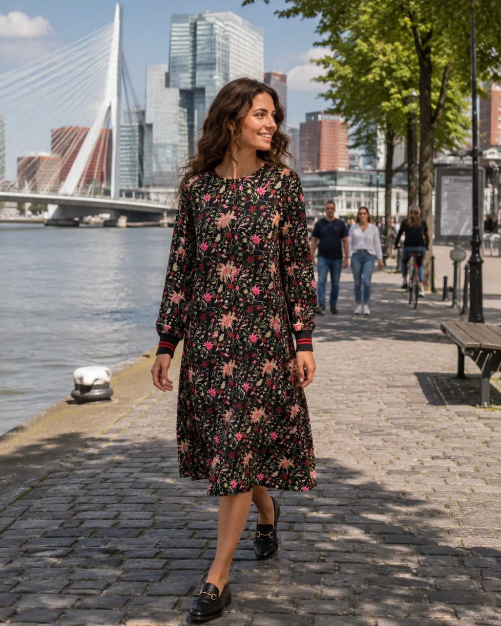 Woman in a floral dress walking along a waterfront promenade with a cityscape in the background.