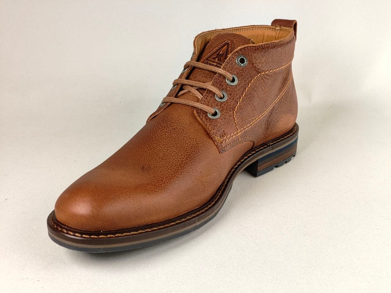 A single brown leather ankle boot with a low stacked heel, brown laces, and metal eyelets, photographed on a light neutral background.