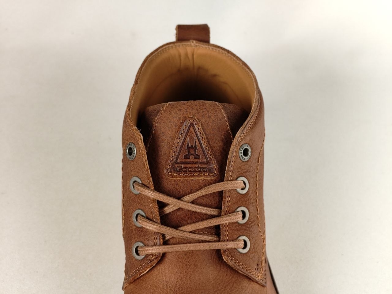 A close-up top-down view of a brown leather lace-up boot, showing the tongue with a triangular logo patch and metal eyelets, on a light neutral background.
