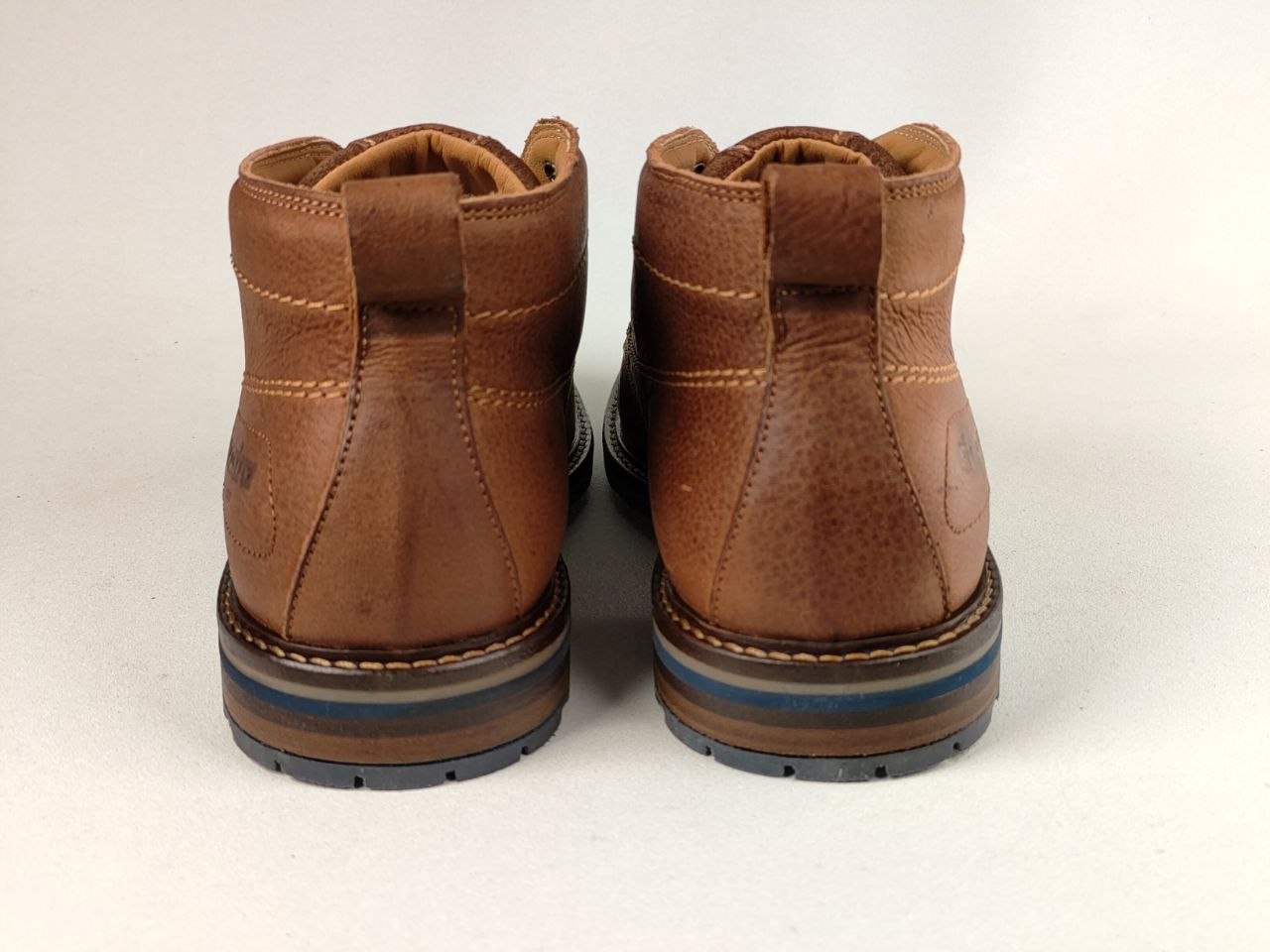 A pair of brown leather ankle boots shown from the back, featuring pull tabs, contrast stitching, and stacked soles with blue and black layers, set against a plain light background.