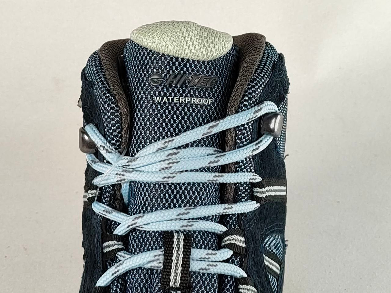 Close-up of the top of a dark blue and gray waterproof hiking boot with light blue laces, on a light beige background.