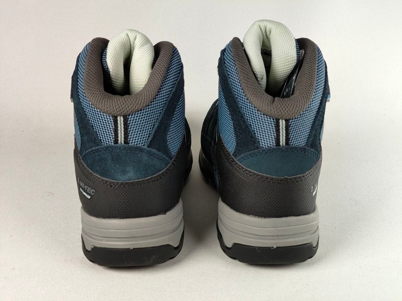 A pair of mid-cut hiking boots shown from the back, featuring blue and gray mesh and suede uppers, dark gray soles, and off-white tongue lining, photographed against a neutral light gray background.