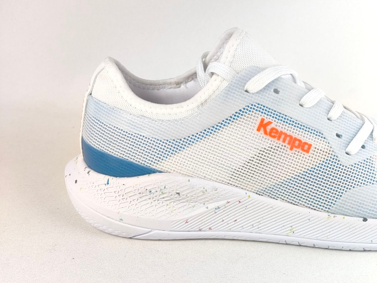 A white Kempa athletic shoe with light blue mesh panels, a blue heel accent, and an orange Kempa logo on the side, shown against a light gray background.