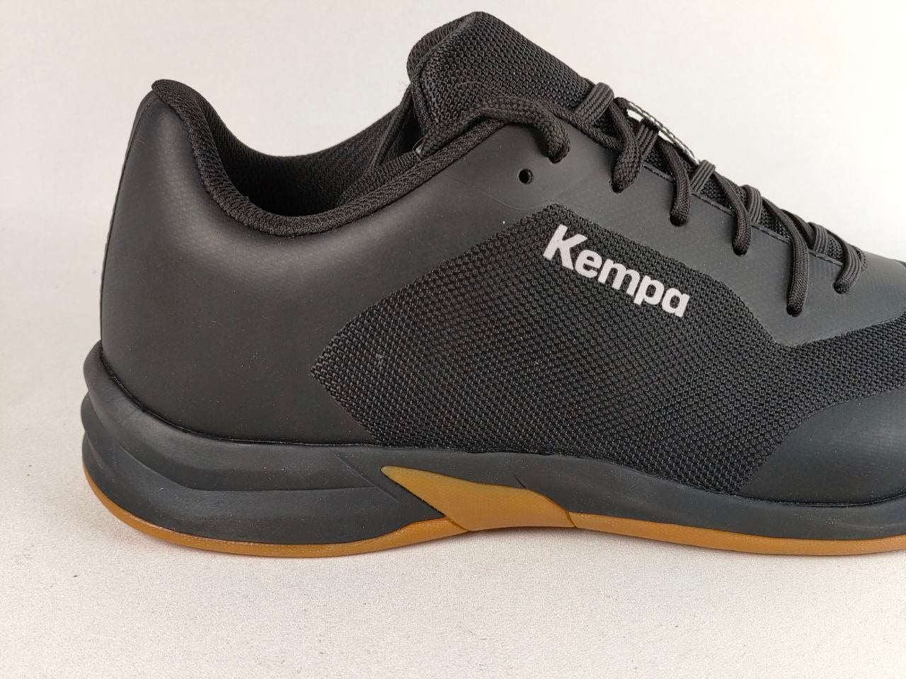 A black Kempa athletic shoe shown in a side profile view against a light gray background. The shoe features a black mesh and synthetic upper with the Kempa logo printed in white on the side, a black sole, and a contrasting brown outsole.