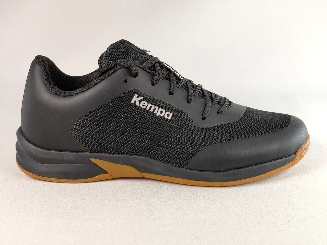 A black Kempa athletic shoe with a textured mesh upper, black laces, and a gum-colored outsole, shown from a side profile on a light gray background.