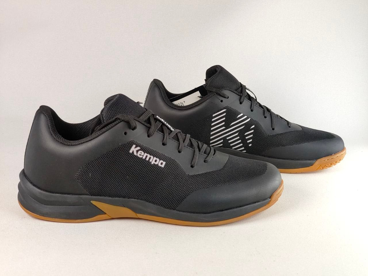 A pair of black Kempa indoor court shoes with gum brown soles and white logo details, photographed on a light gray studio background.