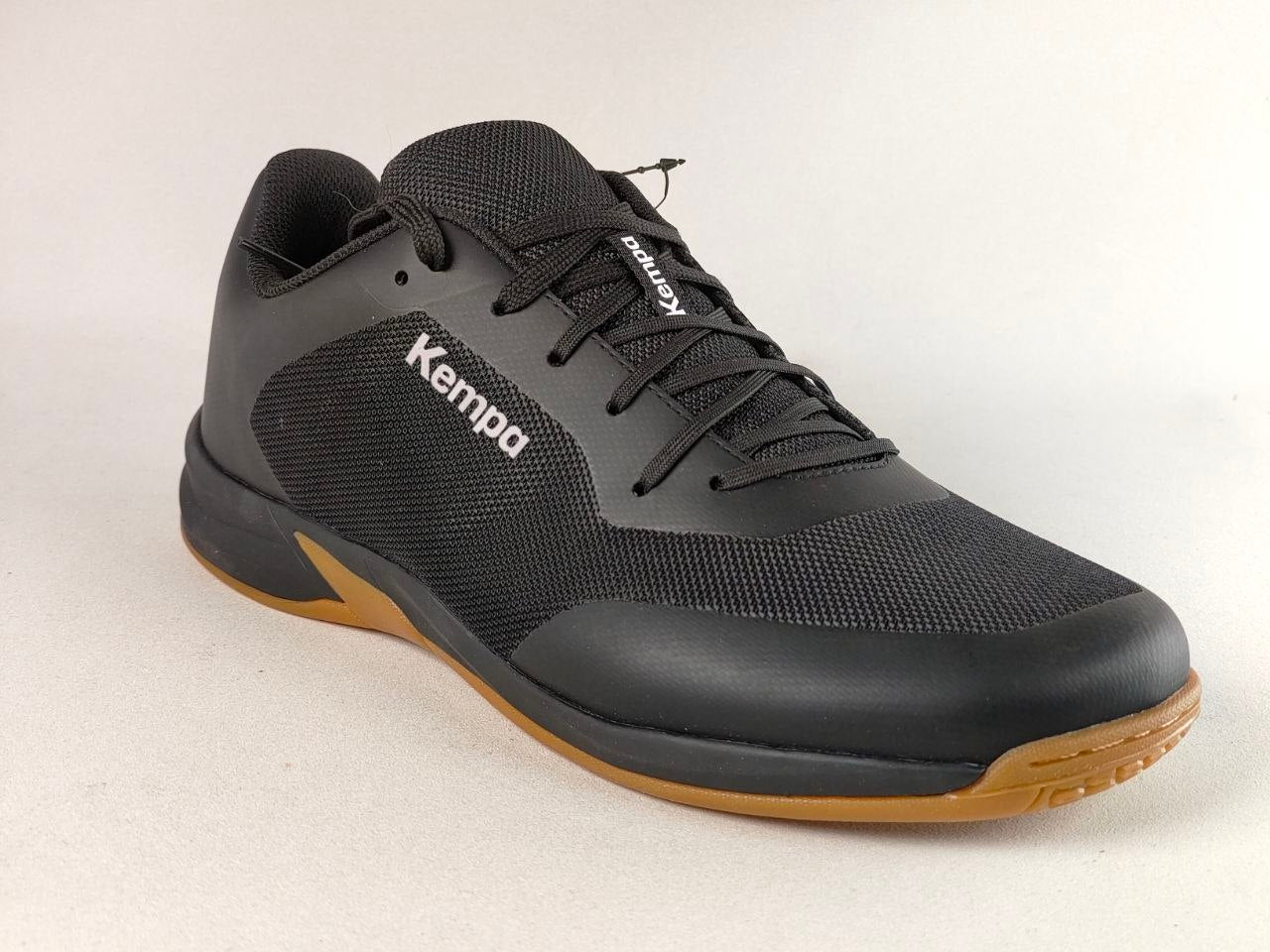 A black Kempa athletic shoe with a textured mesh upper, black laces, and a contrasting brown gum sole, shown against a light gray background.
