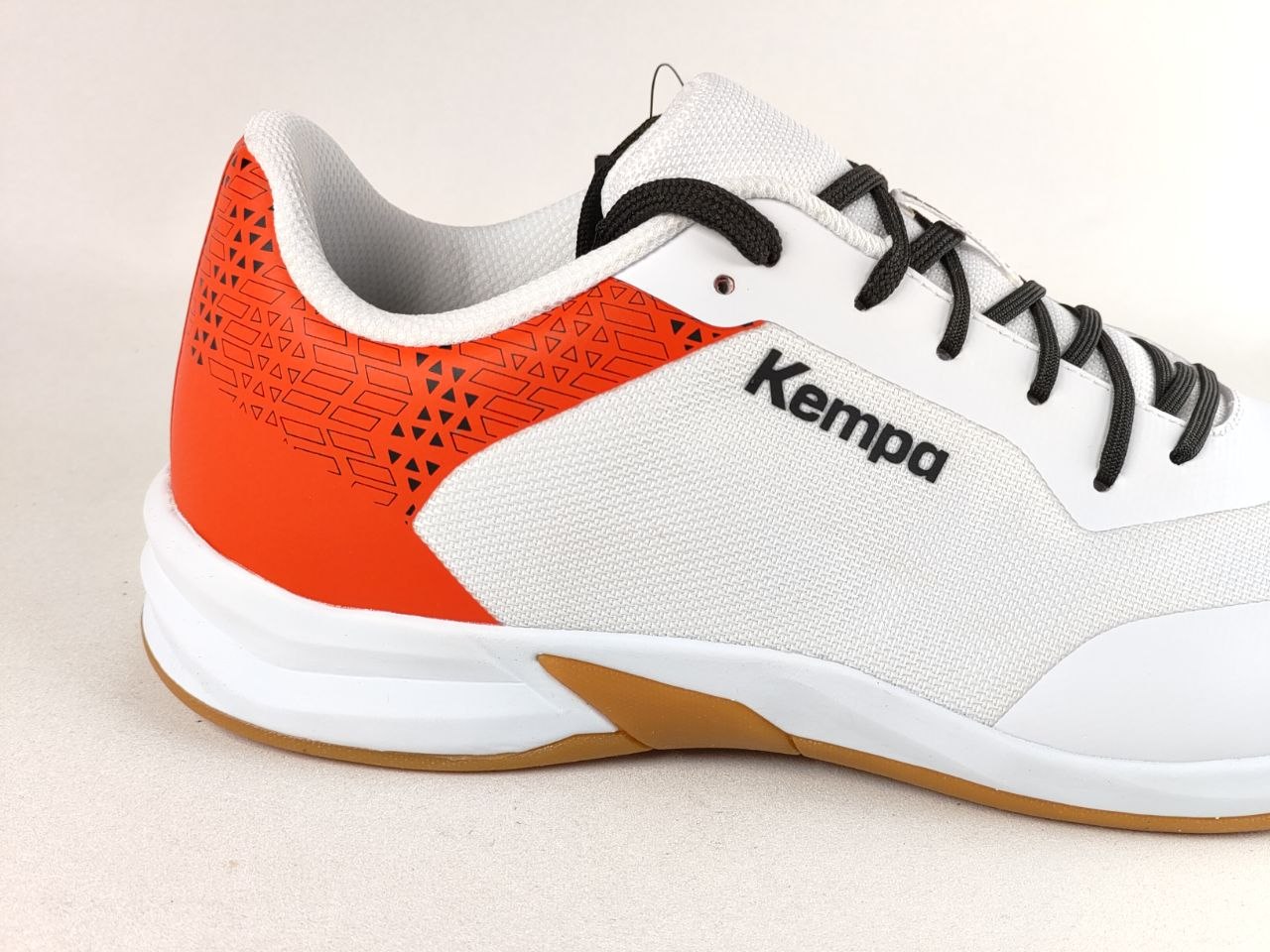 A close-up side view of a Kempa athletic shoe in white, bright orange, and black, with a white and brown sole, set against a light beige background.