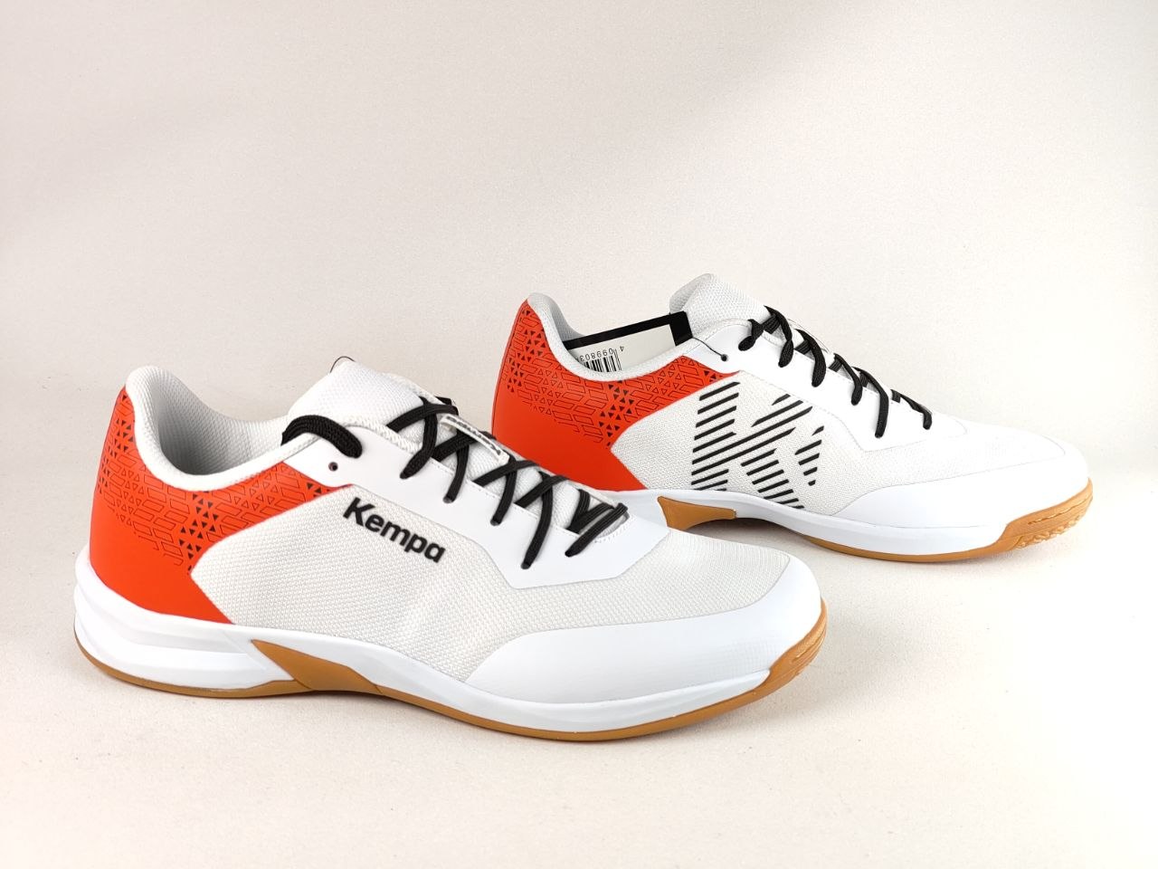A pair of white and orange Kempa athletic sneakers with black laces and a gum sole, displayed on a light beige background.
