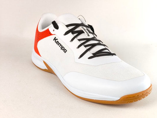 A white Kempa indoor court shoe with black laces, a bright orange-red heel accent, and a gum brown outsole, shown on a light beige background.