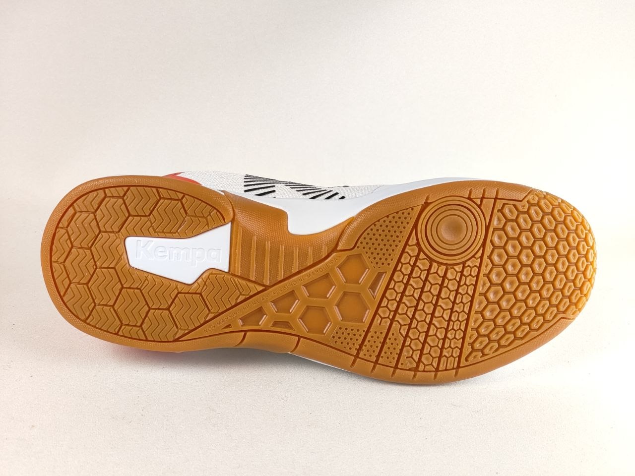 The outsole of a Kempo indoor soccer shoe, featuring a white upper with black graphic stripes and a prominent orange gum rubber sole with textured grip patterns.
