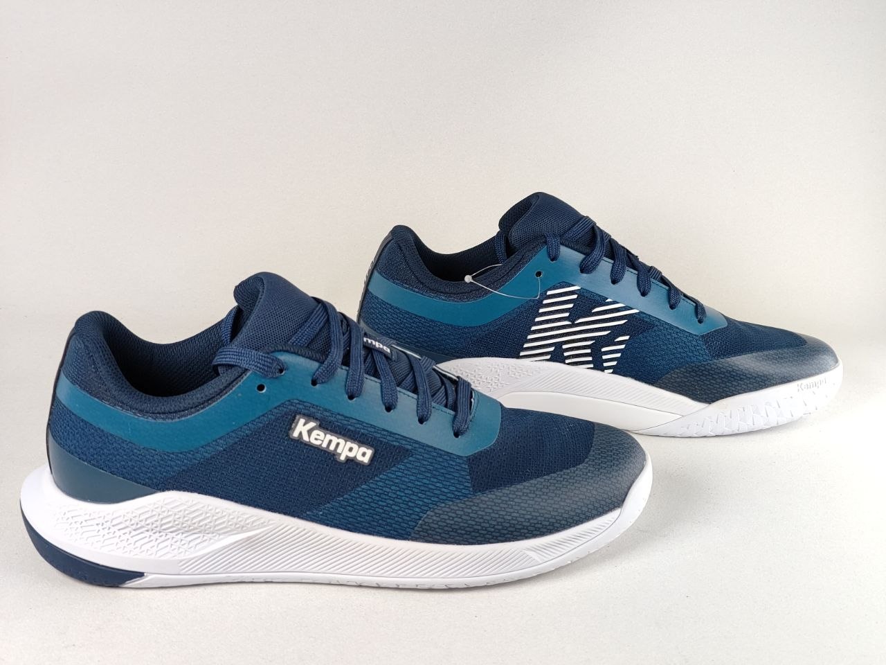 A pair of Kempa athletic sneakers in navy blue and teal, featuring a white midsole and a stylized white 'K' logo on the side, set against a plain light gray background.