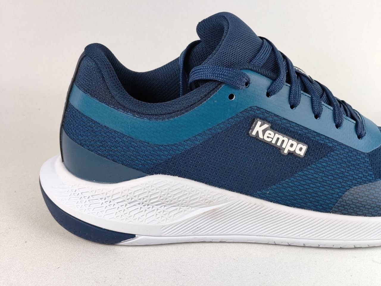 A close-up side view of a Kempa athletic shoe in navy blue and teal with a white midsole, on a light gray background.