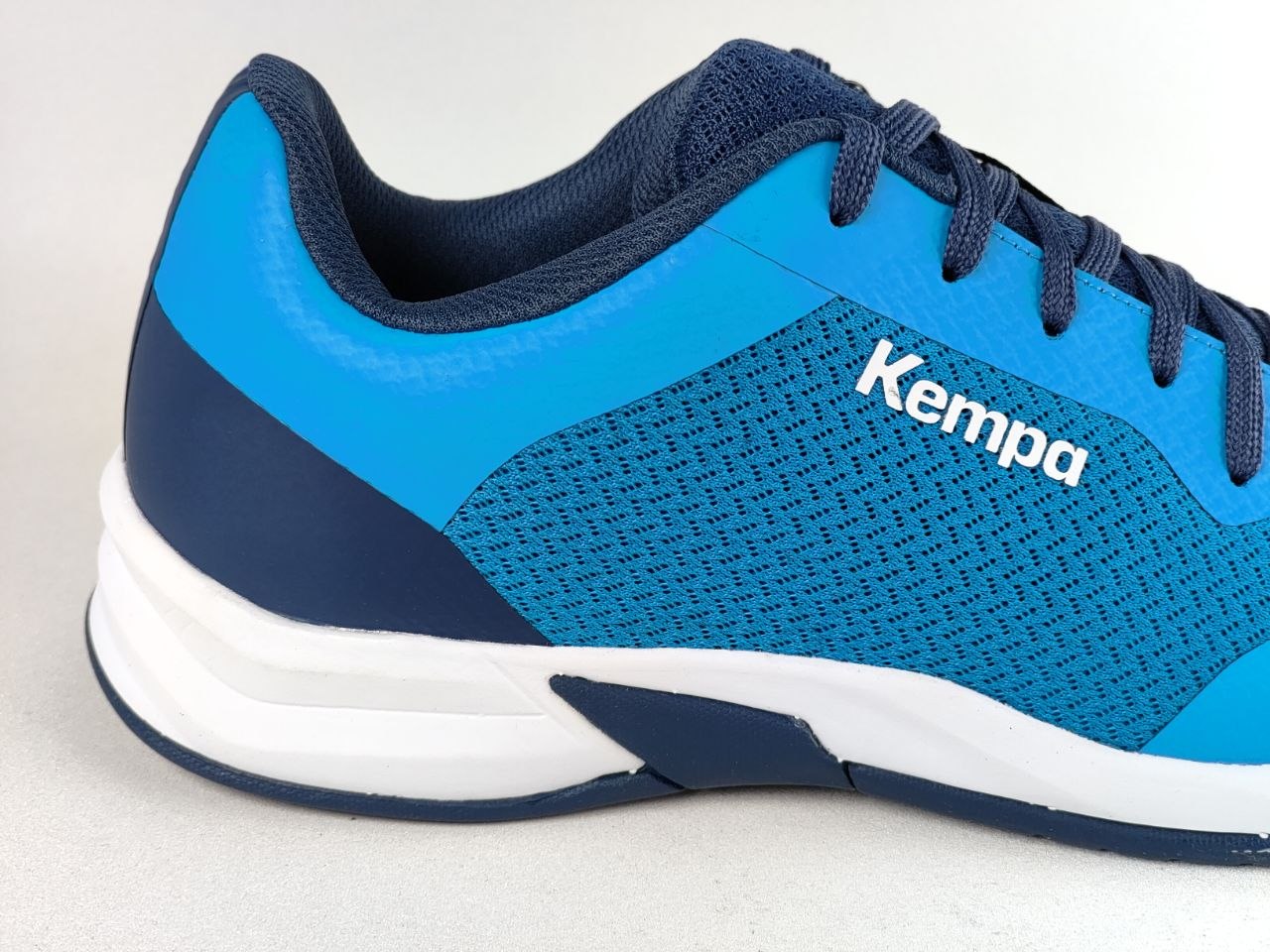 A close-up side view of a blue and navy athletic shoe with the white 'Kempa' logo on the side, set against a light gray background.
