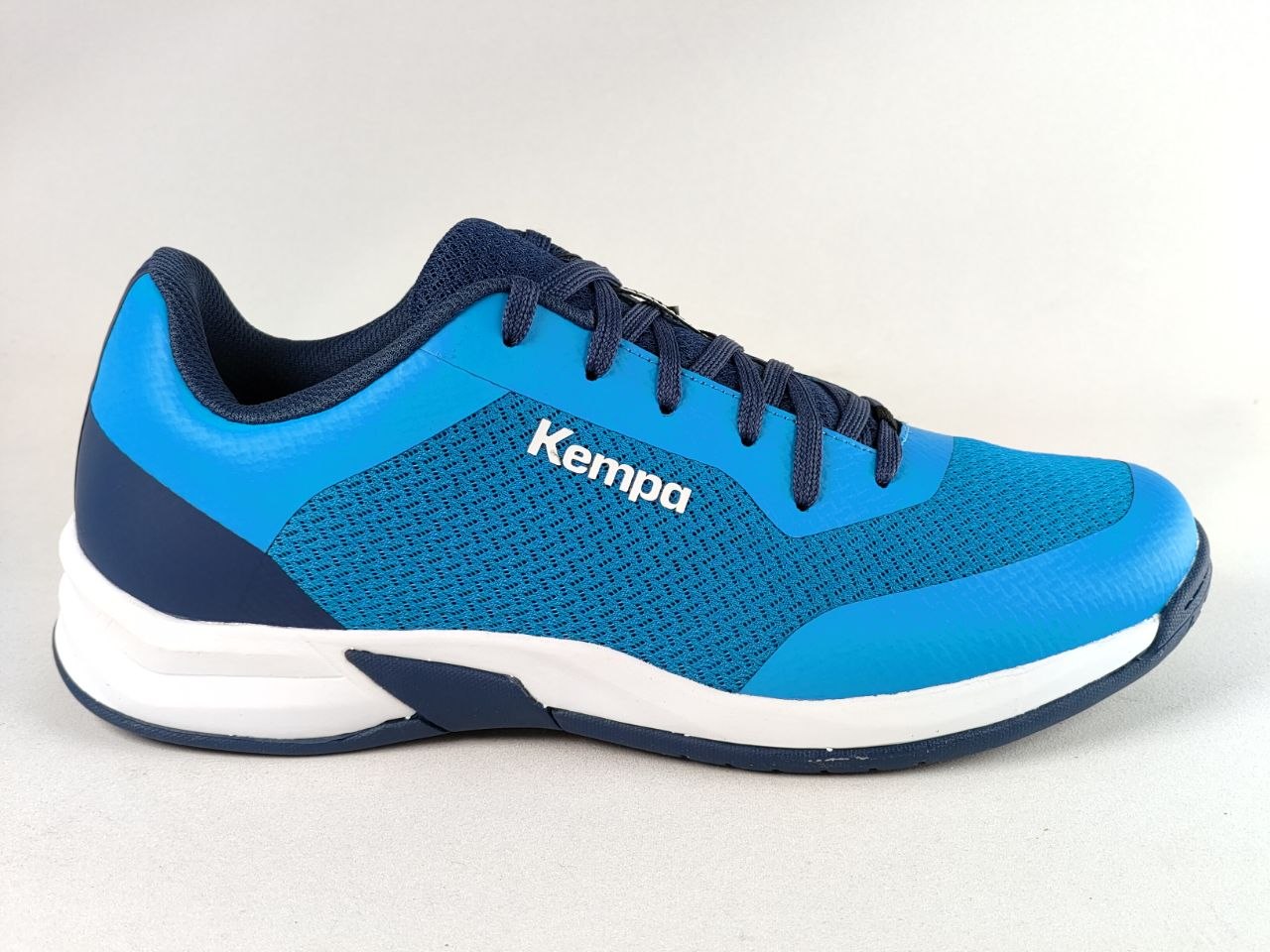A blue and navy athletic shoe with a white midsole, featuring the Kempa logo on the side, photographed against a light gray background.