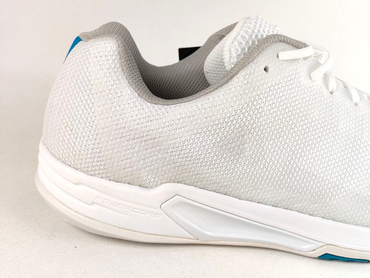 A close-up side view of a white athletic sneaker with a textured knit upper, white sole, and subtle blue accents on the heel and toe.