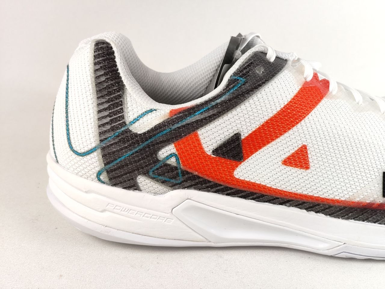 A close-up side view of a white athletic sneaker with a white midsole, featuring a textured black and gray heel panel, a bold orange side stripe, and teal accent lines on a light gray background.