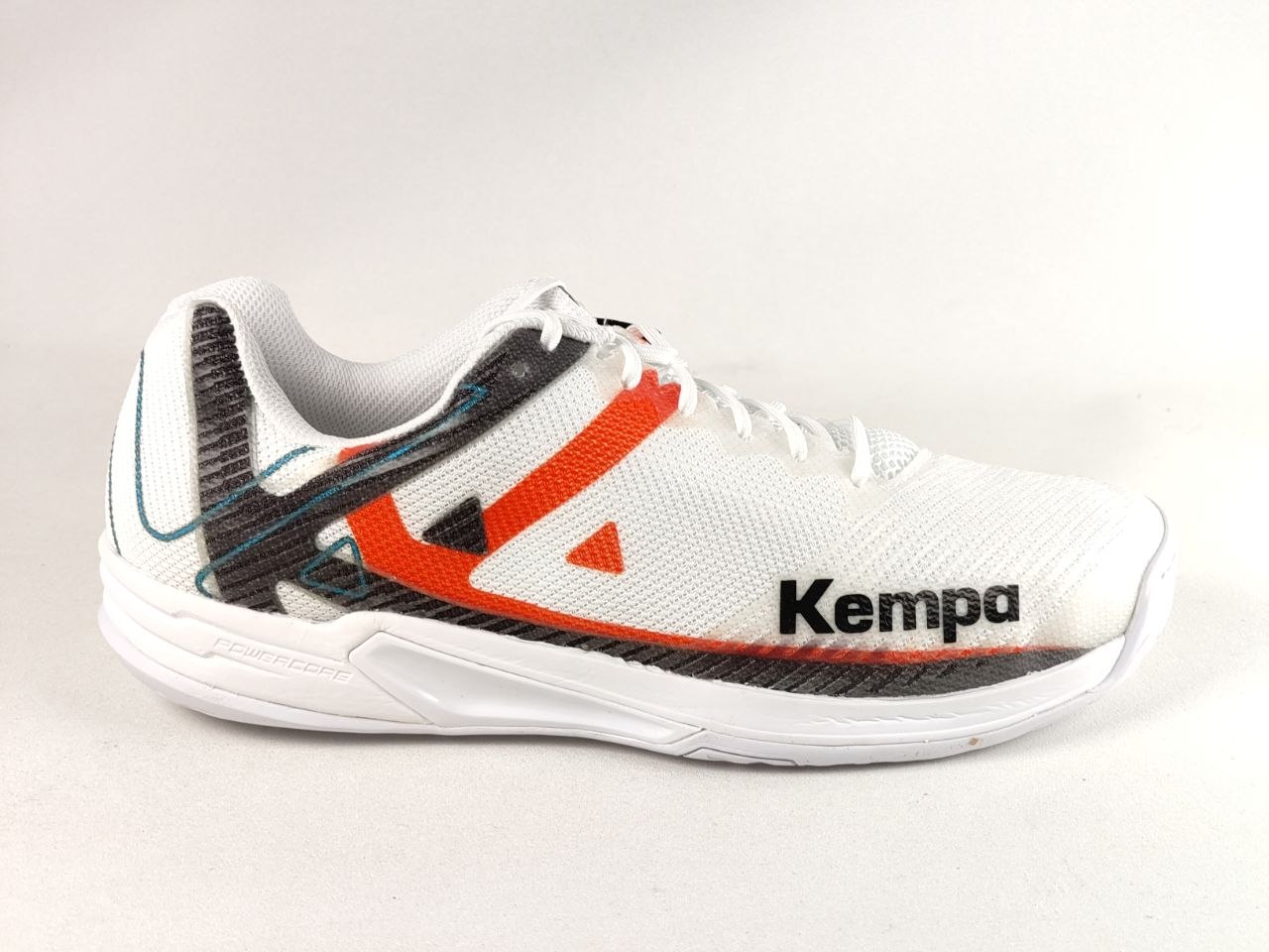 A white Kempa athletic shoe with a white mesh upper, featuring a prominent orange-red side stripe, a black textured heel accent, and the black 'Kempa' brand name on the side, set against a plain light background.