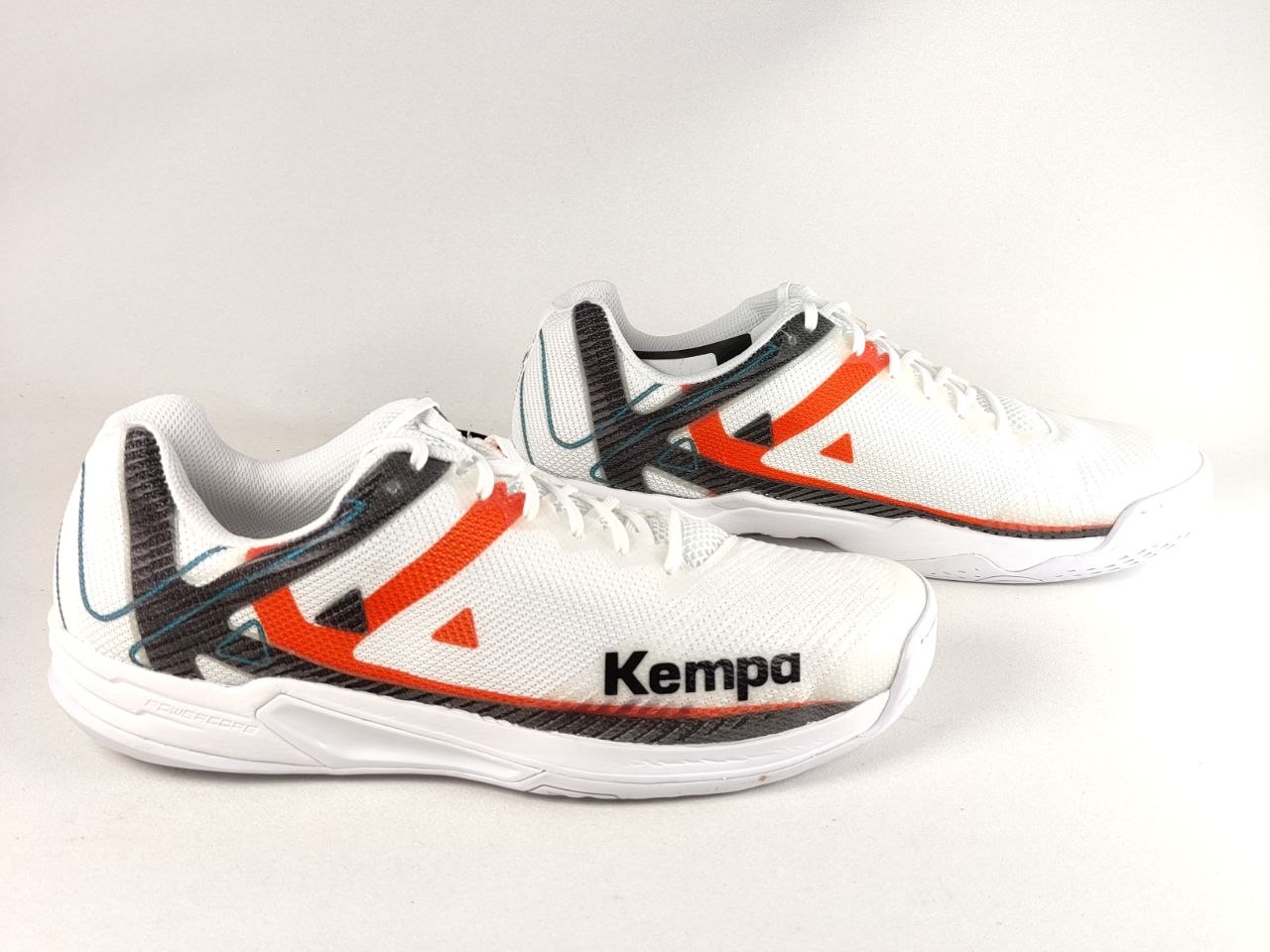 A pair of white Kempa athletic shoes with black and orange accents, displayed on a light gray background.