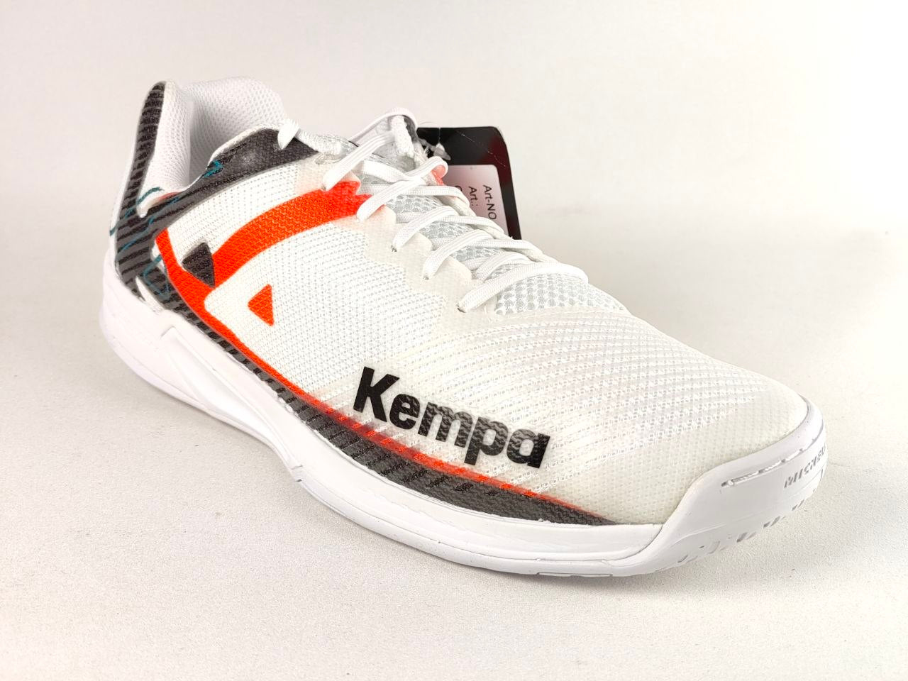 A white Kempa athletic shoe with black and orange accents, featuring a white mesh upper, white laces, and a white sole, photographed on a light beige background.