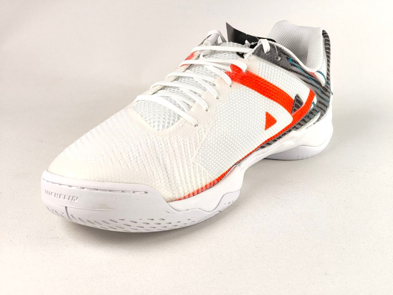 A white athletic shoe with bright orange accents and a textured knit upper, photographed on a light gray background.