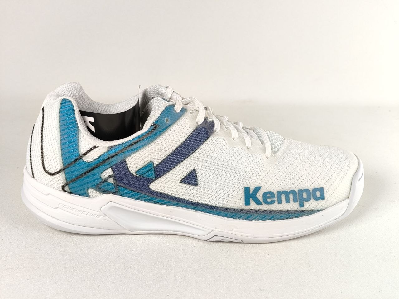 A white Kempa athletic shoe with teal and navy blue accents, shown from a side profile against a light beige background.