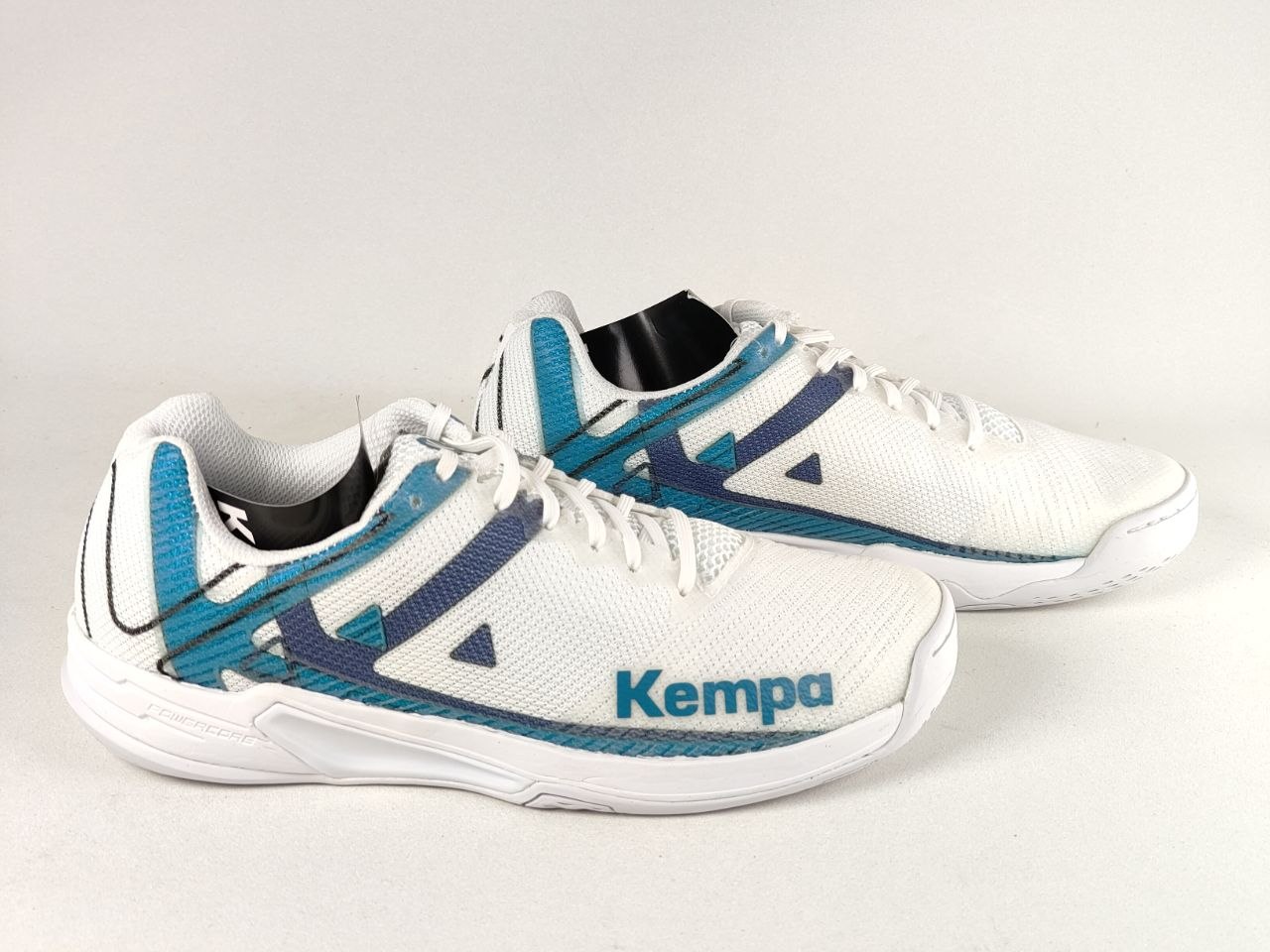 A pair of white Kempa athletic shoes with teal and navy blue accents, displayed on a light gray background.
