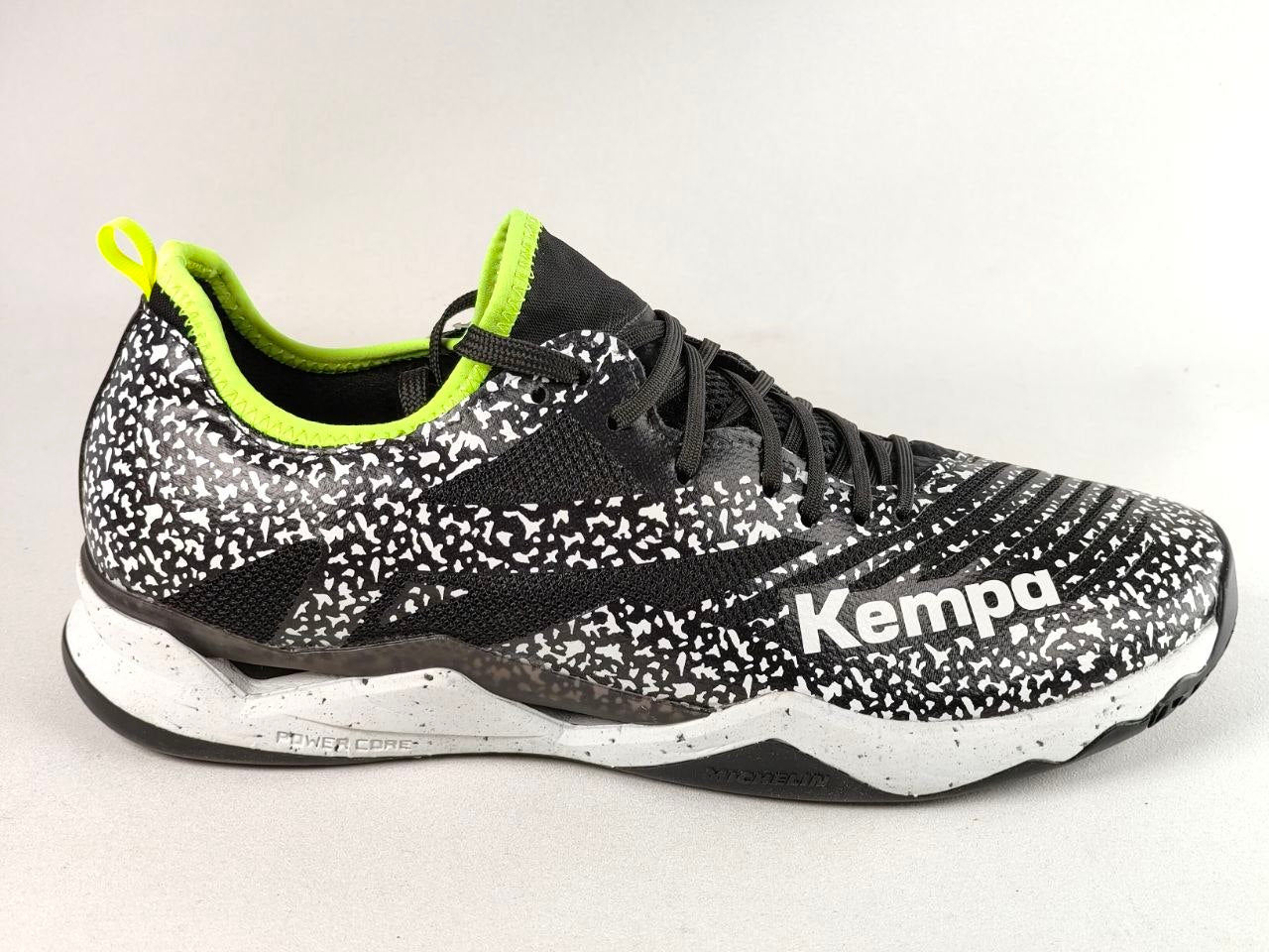 A single Kempa athletic shoe shown in a side profile view against a light gray background. The shoe features a black and white speckled pattern on the upper, black laces, a bright neon yellow-green collar lining and pull tab, and a white midsole with black detailing.