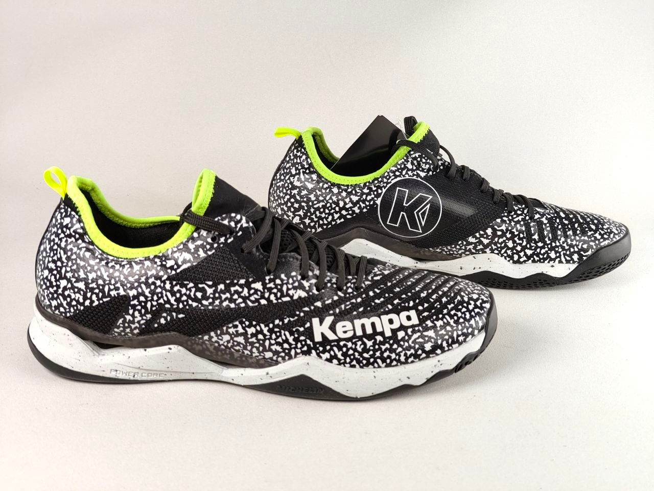 A pair of black and white speckled Kempa athletic sneakers with bright neon yellow-green accents on the collar lining and heel pull tabs, displayed on a light gray background.