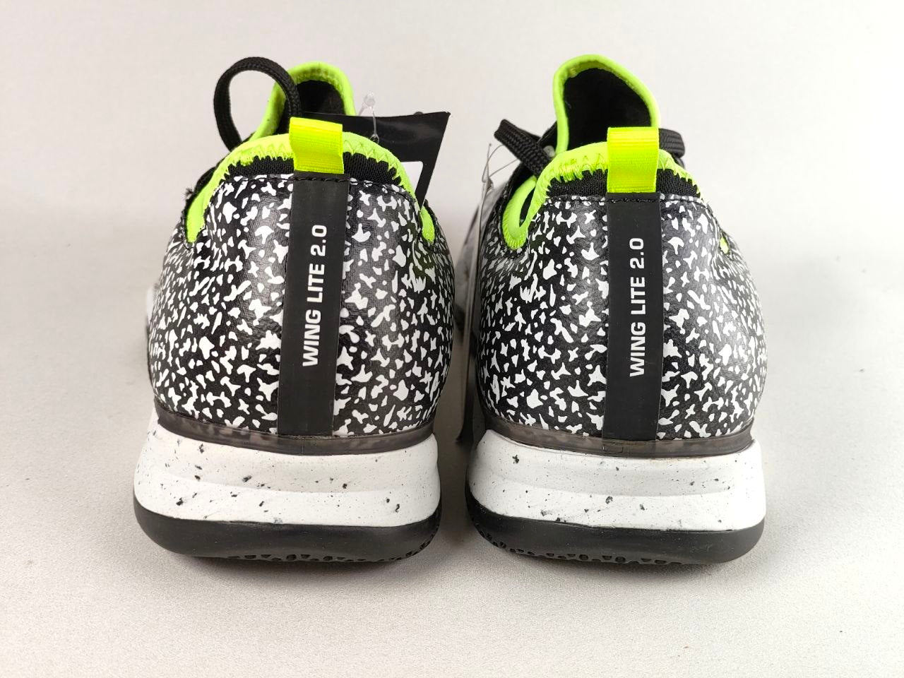 A pair of Nike Wing Lite 2.0 sneakers shown from the back, featuring a black and white speckled pattern, a black heel tab with white text, and a bright neon yellow-green pull tab and lining.
