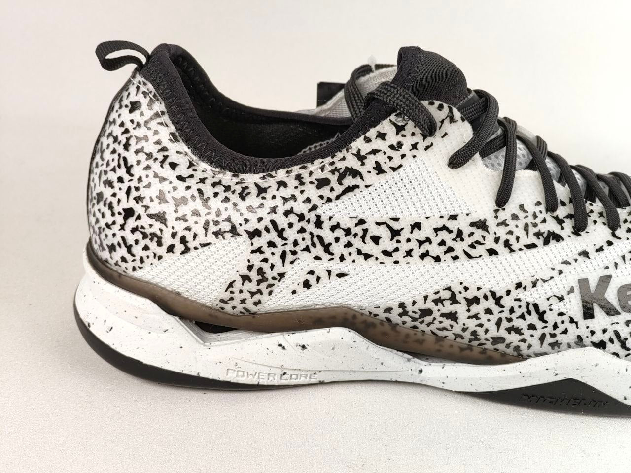 A close-up side view of a Reebok athletic shoe featuring a white and black speckled pattern on the upper, black laces, and a white midsole with a brown accent.