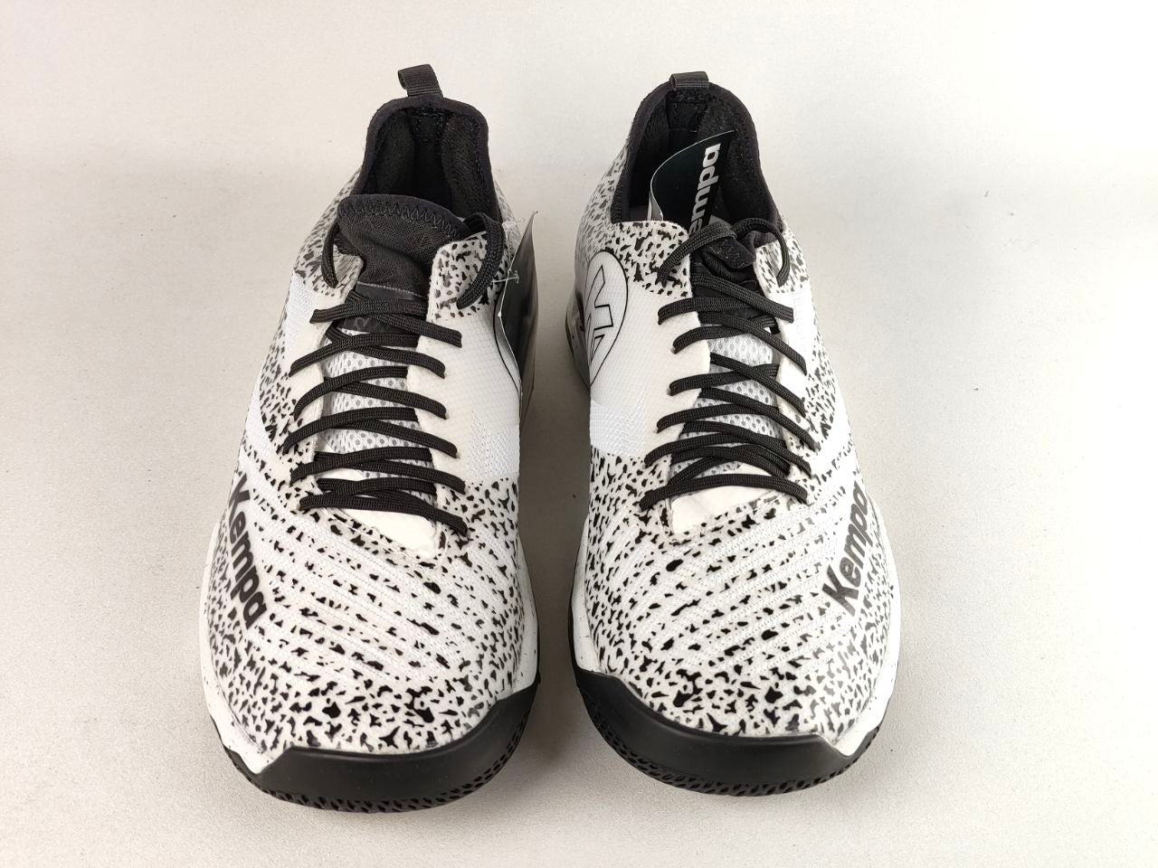 A pair of white and black speckled athletic sneakers with black laces, photographed from the front on a light gray background.