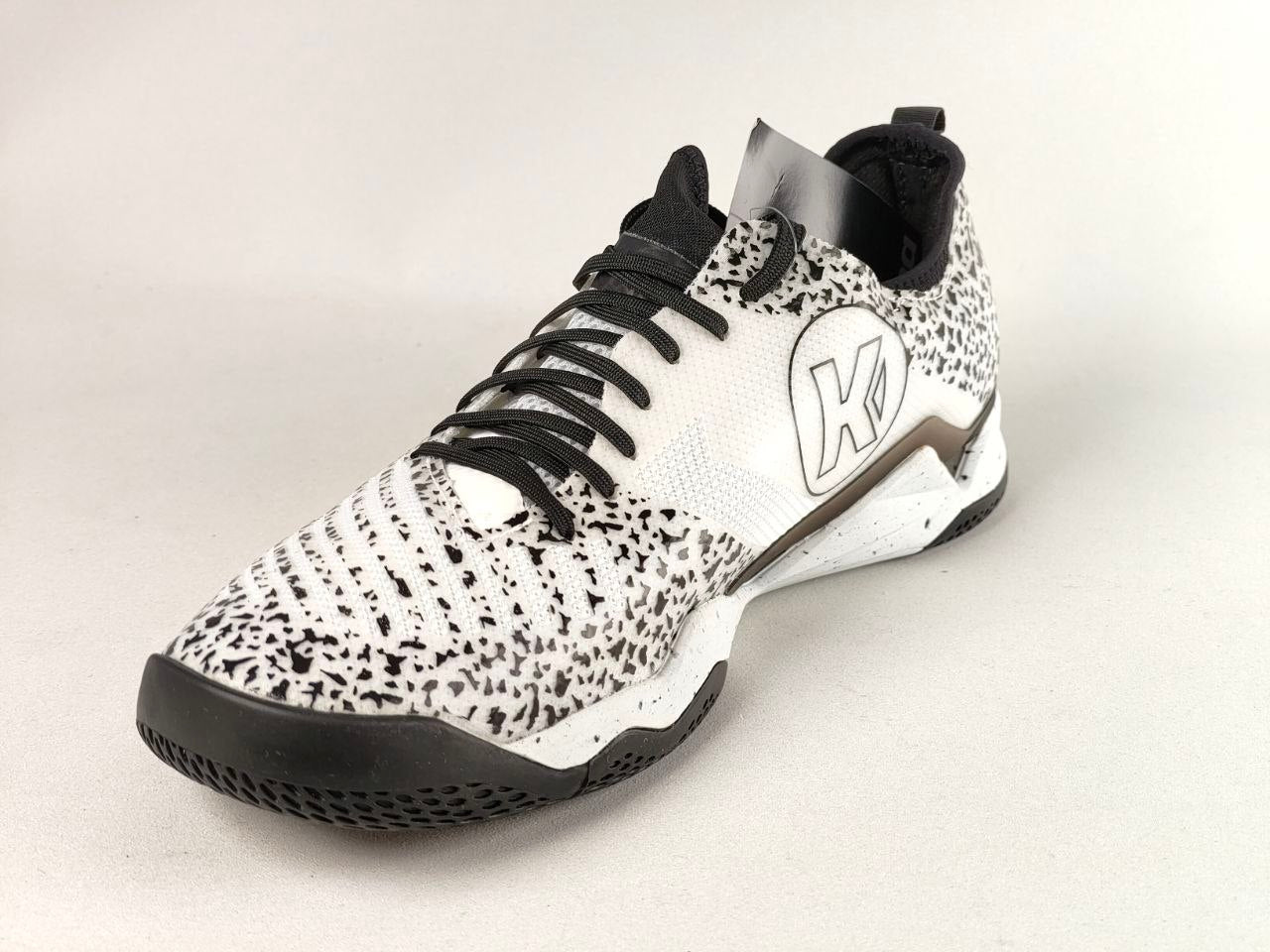 A single athletic sneaker shown at a three-quarter angle on a light gray background. The shoe features a white textured upper covered in black speckled patterning, black laces, a black outsole, and a white midsole with a stylized 'K' logo and a metallic bronze accent.