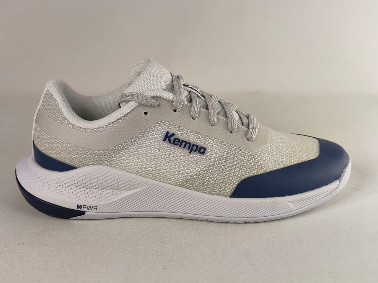 A light gray and white Kempa running shoe with navy blue accents on the toe cap and midsole, shown from a side profile against a neutral light gray background.
