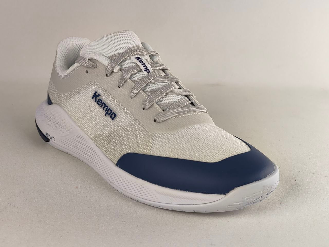 A low-top athletic sneaker by Kempa, featuring a light gray and white mesh upper with a navy blue toe cap and matching navy branding on the side, set against a neutral light gray background.