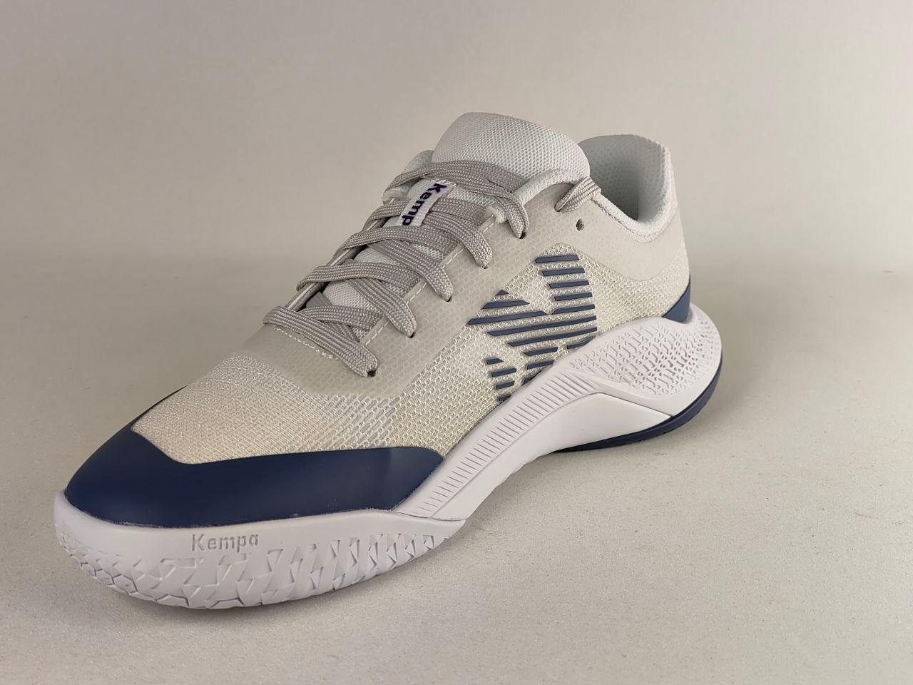 A single athletic sneaker shown from a three-quarters angle on a neutral light gray background. The shoe features a light gray mesh upper, a navy blue toe cap, a navy blue side logo, and a white midsole with 'Kempa' branding on the forefoot.