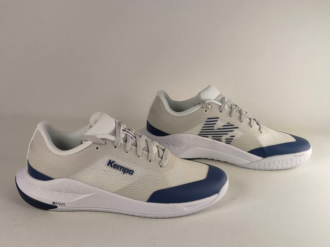A pair of Kempa brand athletic sneakers in off-white and navy blue, displayed on a neutral light gray background.