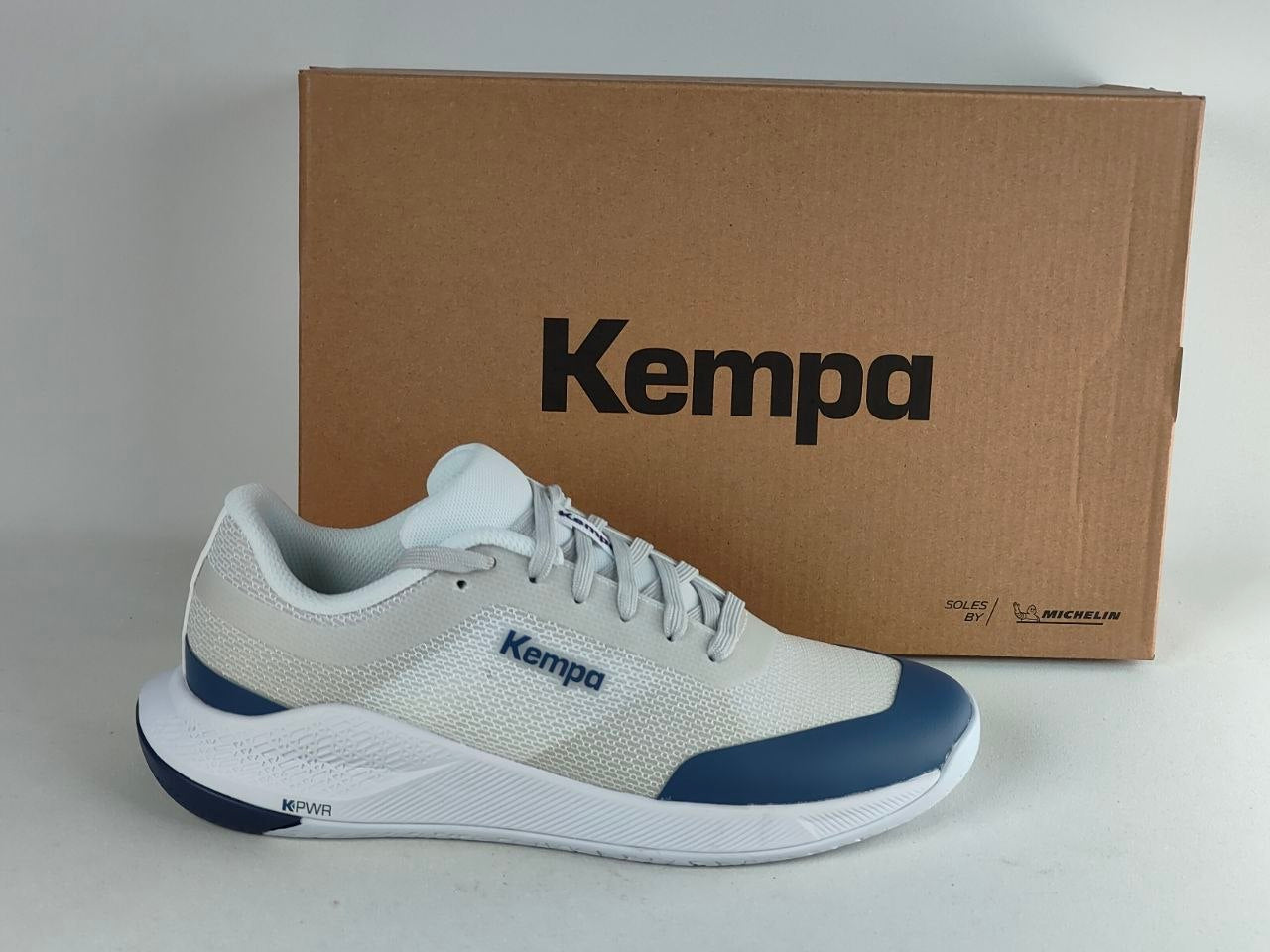 A white and navy blue Kempa running shoe positioned in front of its brown cardboard packaging box on a light gray background.