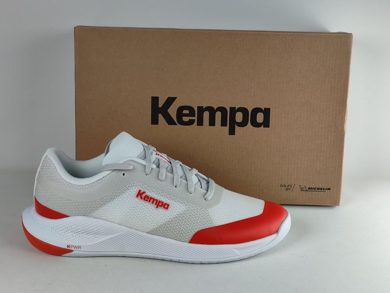 A white and red Kempa running shoe positioned in front of its brown cardboard packaging box on a light gray background.