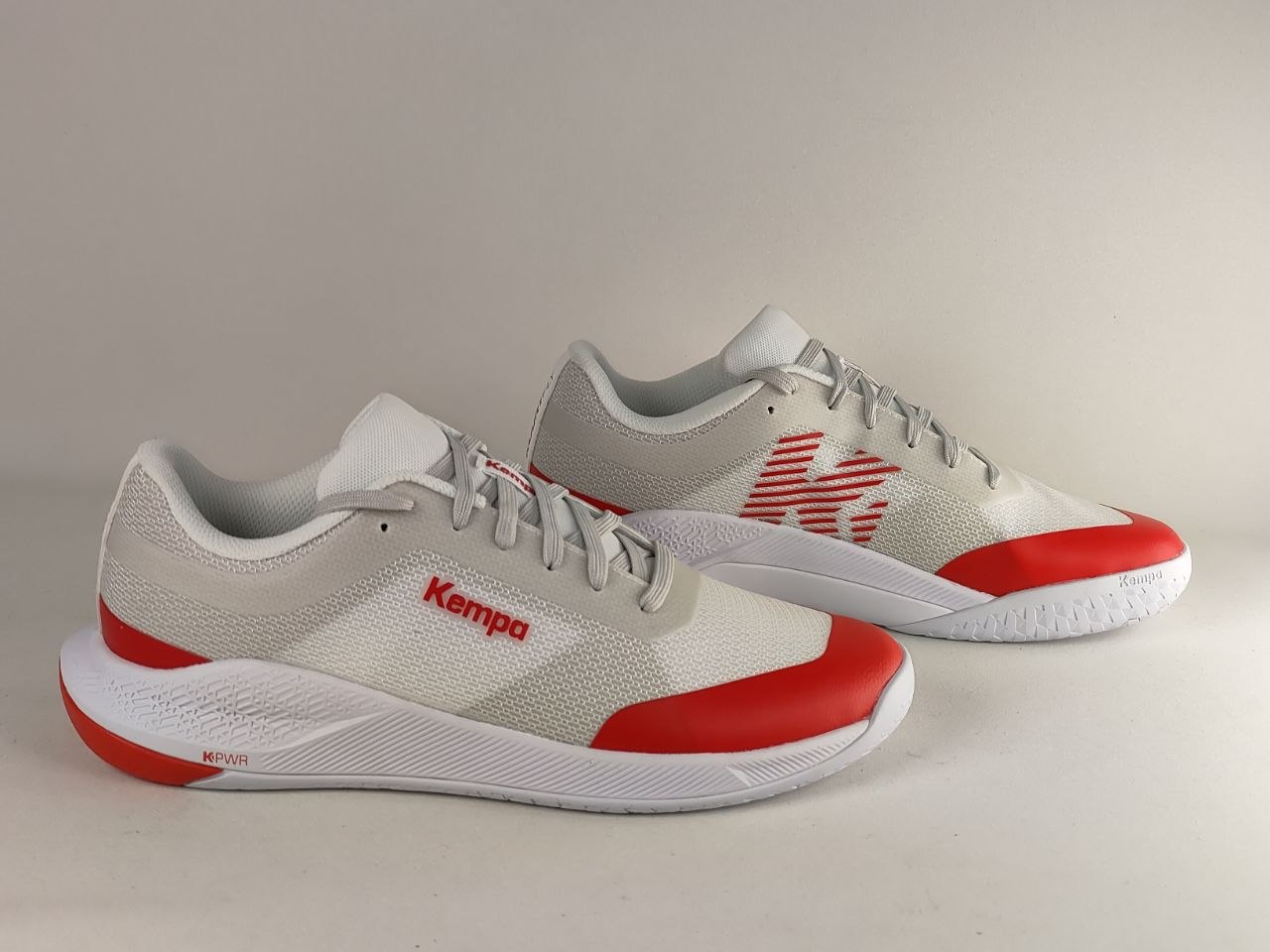 A pair of Kempa brand athletic sneakers in light gray and white with vibrant red accents on the toe caps and side branding, displayed against a neutral light gray background.