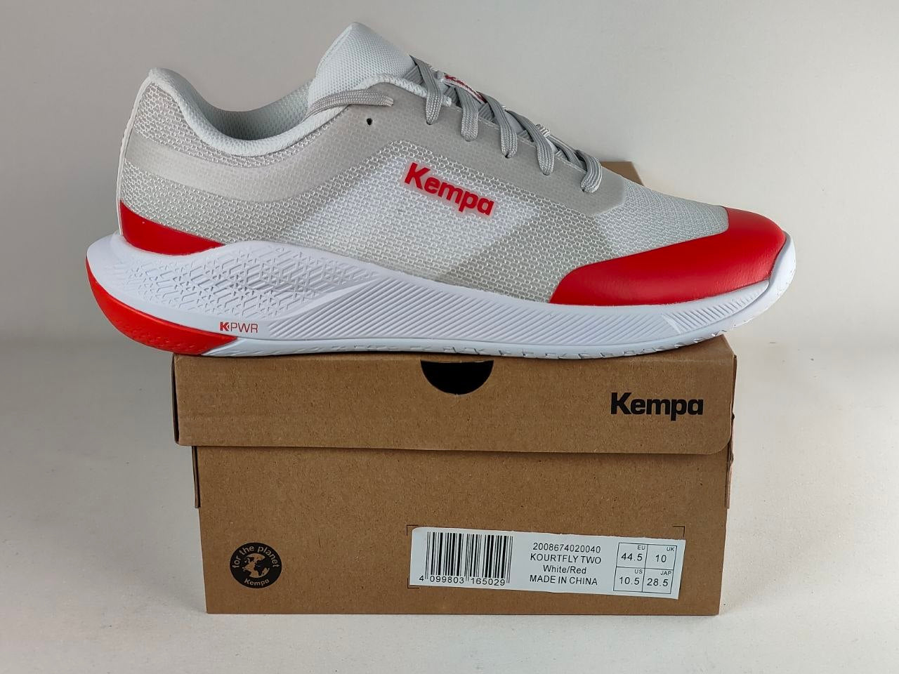 A Kempa athletic sneaker in white and gray with red accents, resting on top of its brown cardboard shoebox against a neutral light gray background.