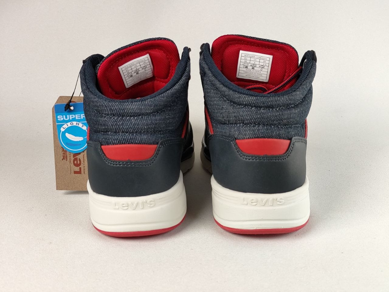 A pair of Levi's high-top sneakers shown from the back, featuring a dark gray denim-like upper, navy heel panels, bright red lining and accents, and white soles. A blue and brown Levi's Super 700 tag is attached to the left shoe on a light gray background.