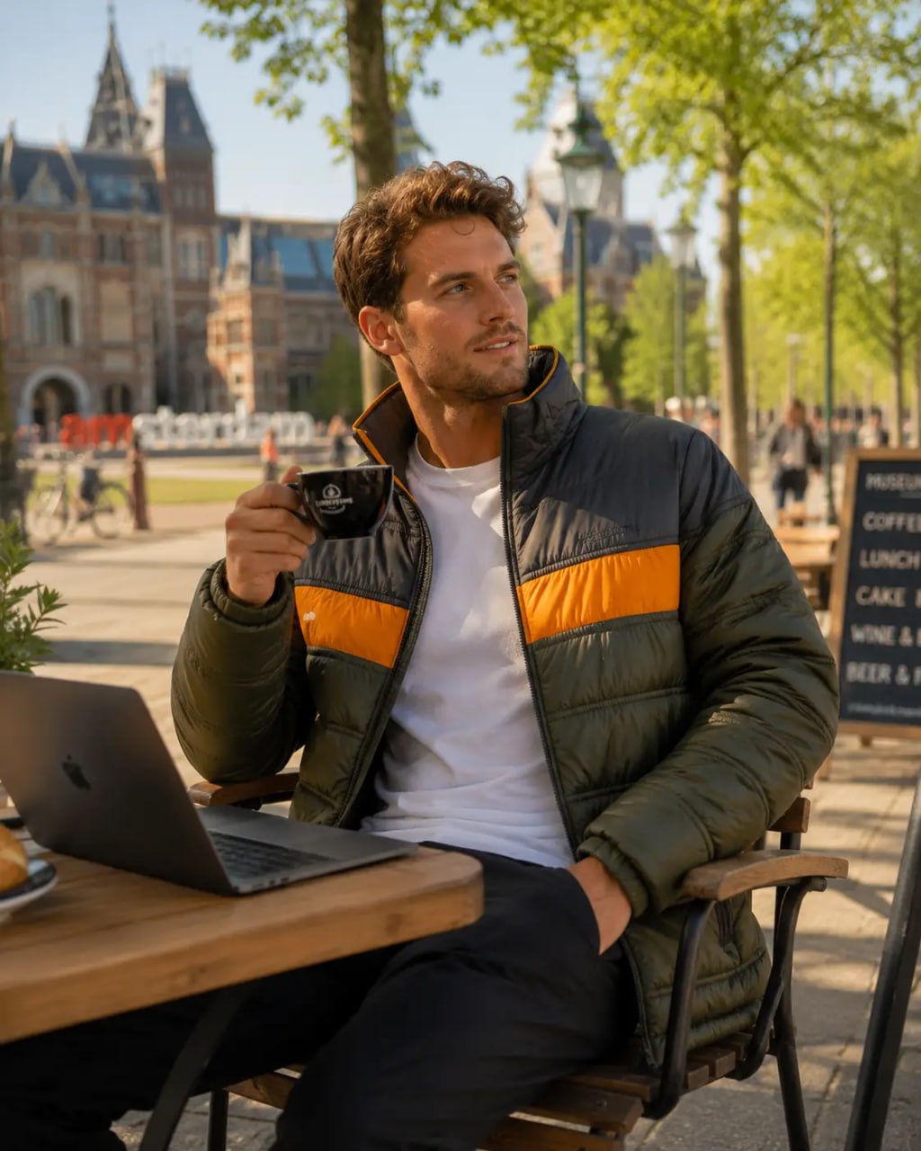 Man sitting outdoors with a laptop and coffee, surrounded by trees and buildings.